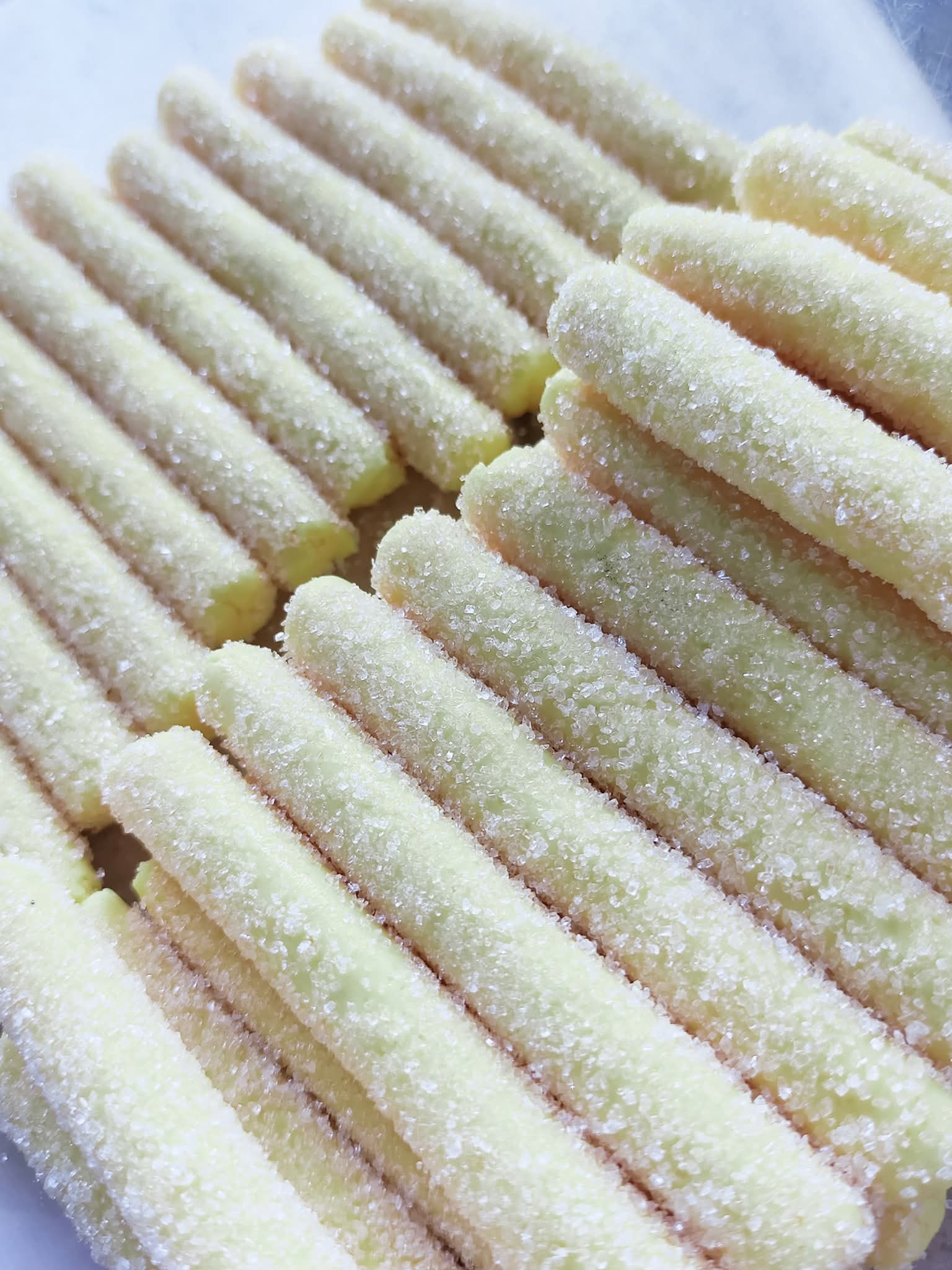 Pale yellow, stick-shaped snacks heavily coated in granulated sugar crystals, arranged in parallel rows.